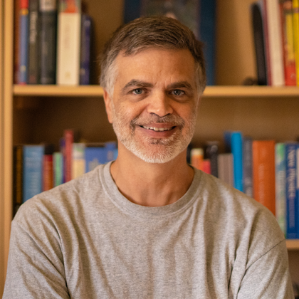 Positive Vive Picture of Erik Larson; a smiling Caucasian man wearing a light-gray T-shirt, seated in front of a bookcase filled with books.